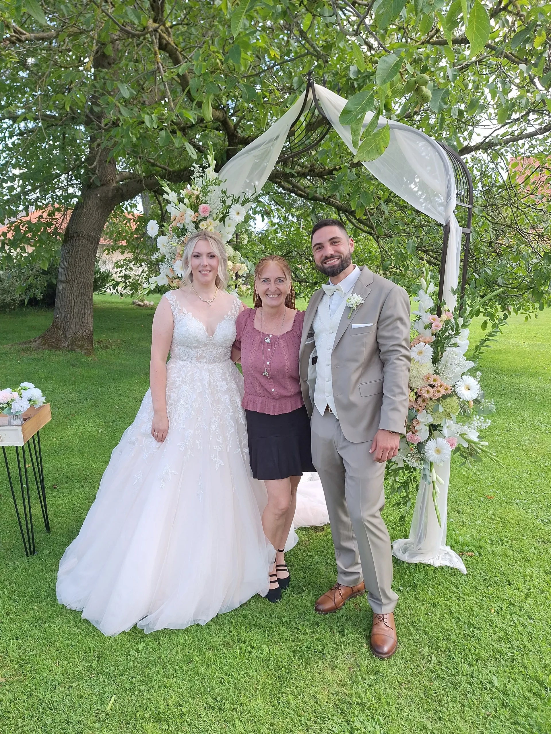 Hochzeitspaar mit Traurednerin auf der Wiese vor einem Traubogen und einem  Baum